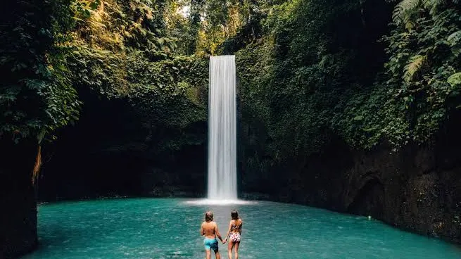 Two visitors standing at the base of Tibumana Waterfall in Ubud Bali with turquoise pool and lush jungle surrounding them