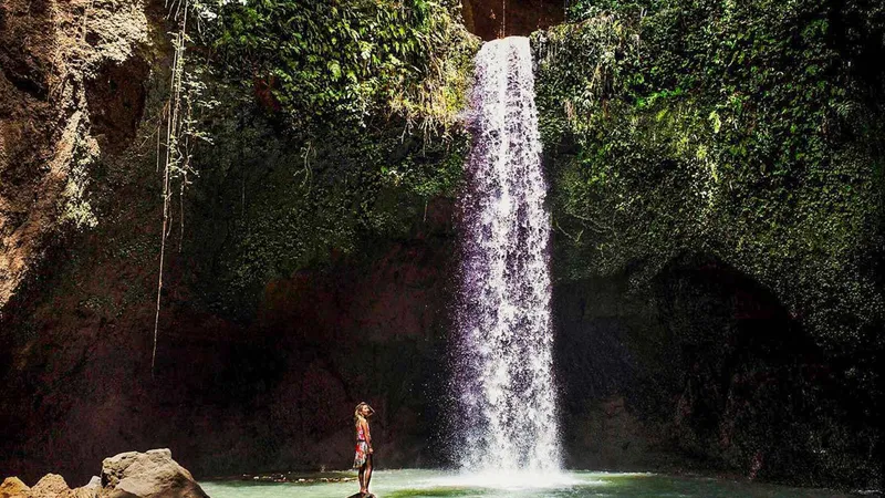 A lone visitor beneath the tall single drop of Tibumana Waterfall near Ubud, one of three stops on a Bali waterfall tour