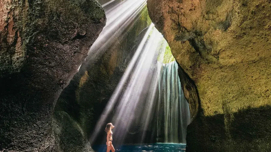 Column of sunlight falling through the cave roof above Tukad Cepung waterfall in Ubud Bali as visitors wade through the shallow canyon river below